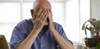 Elderly man sitting at a table with his hands covering his face, surrounded by paperwork